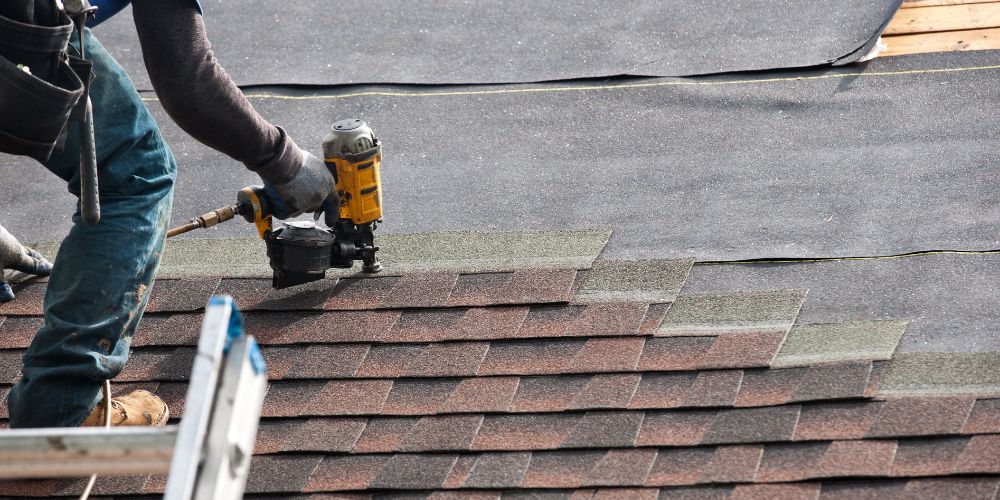 A man off-camera re-shingling a house using a bright yellow power nail gun, and dark brown shingles with slight reddish undertones.