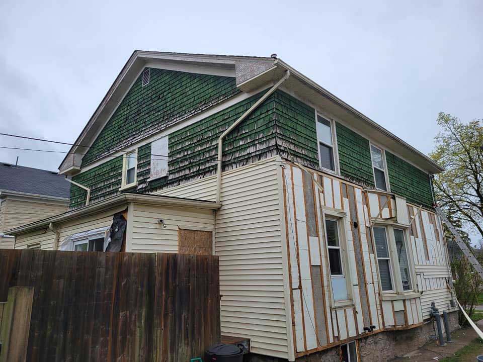 the image shows a home siding replacement being done by our Quality Care Roofing team, the top half of the home has decaying siding panels while the bottom half has brand new cream panels.